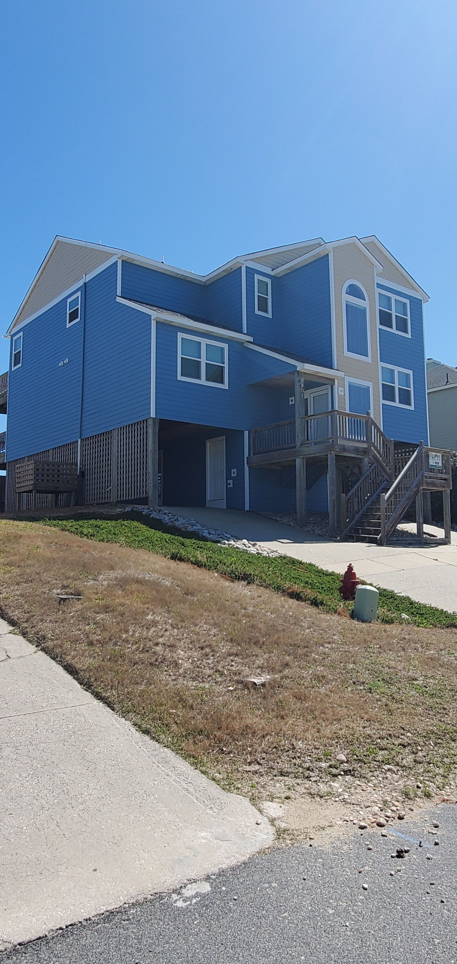 Blue elevated house on stilts with stairs, beside a sloped driveway under a clear sky
