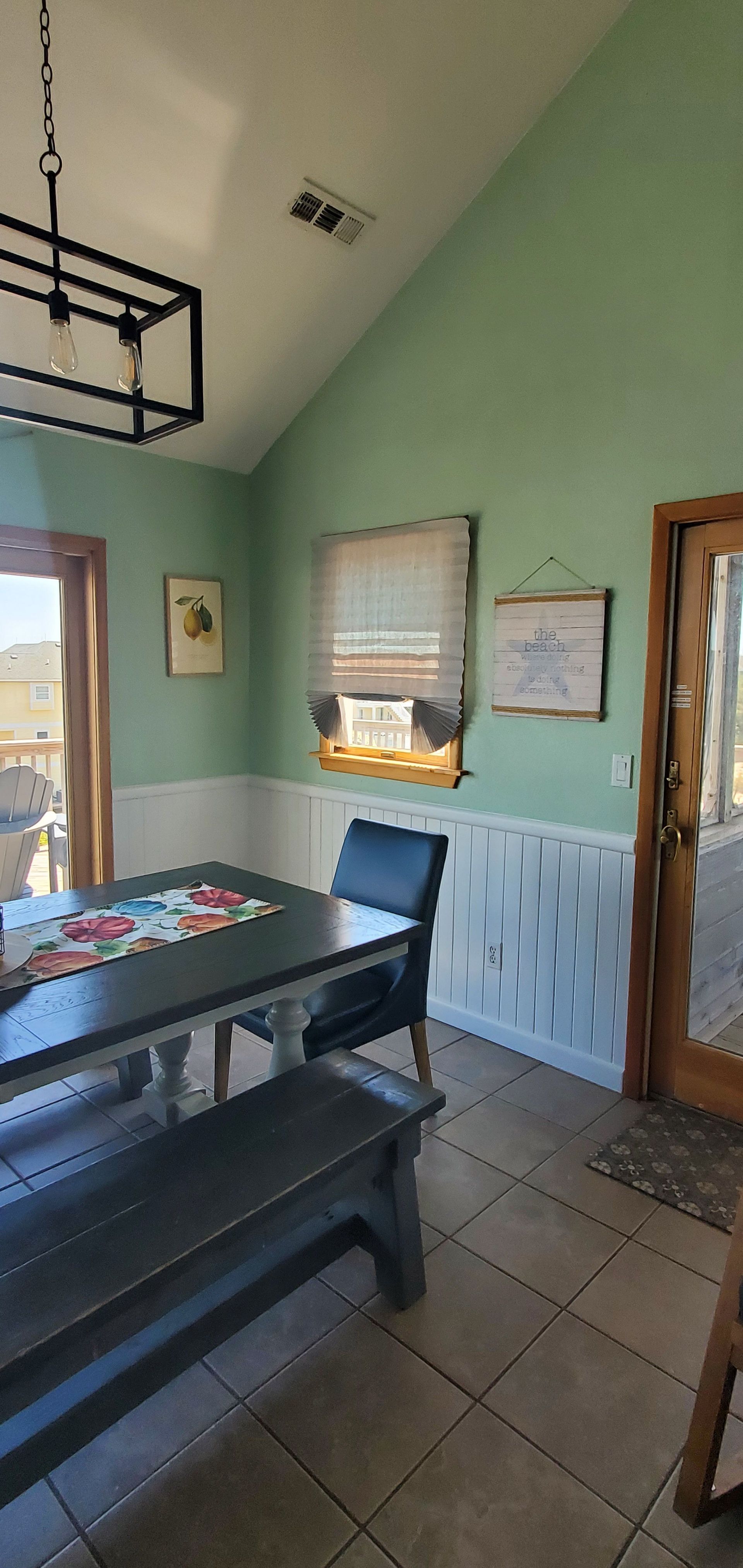 Dining room with green walls, tiled floor, black table and bench, and a window letting in sunlight