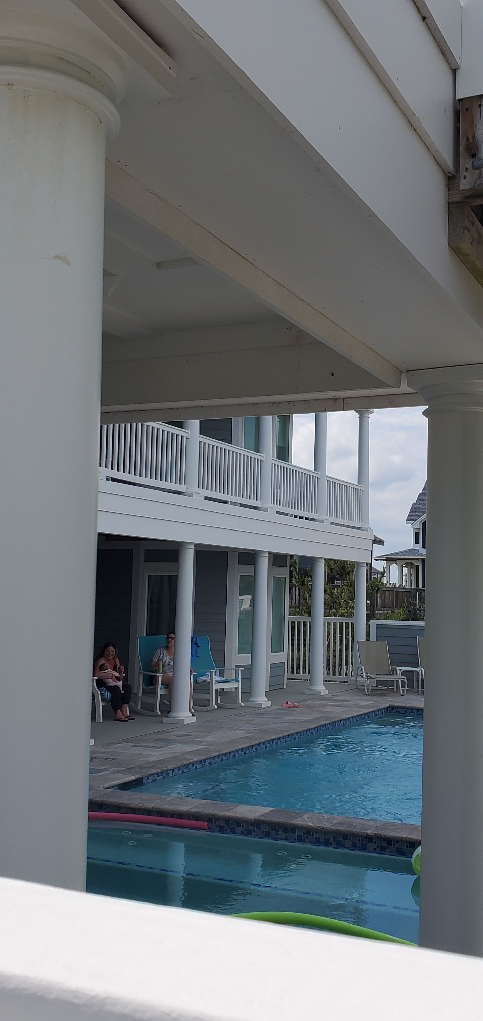 Covered outdoor pool with white columns and a building walkway above, with people seated by the water.