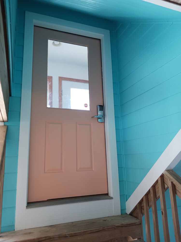 Pastel pink door in a blue entryway with a small window and wooden railing.