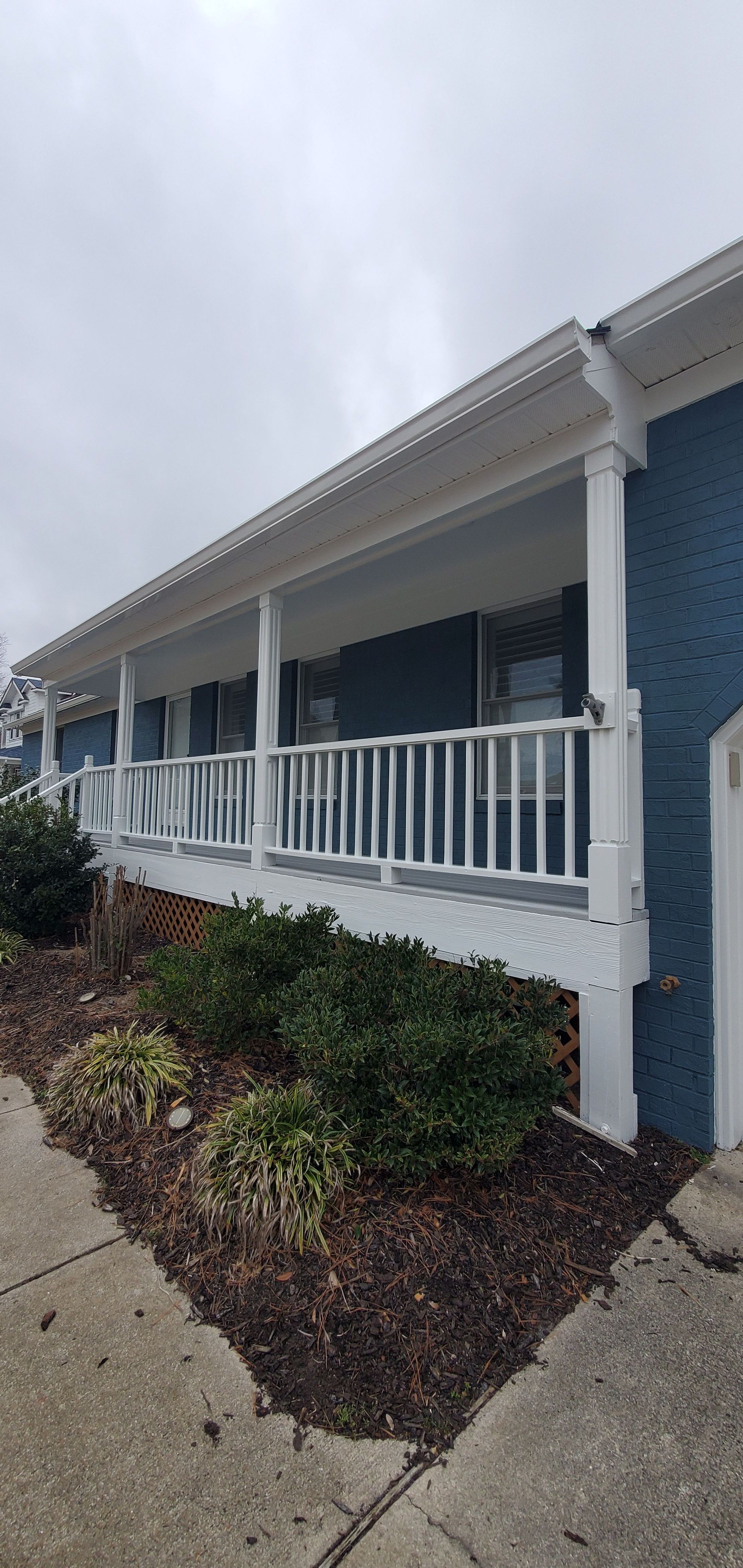 White porch with railings and blue siding on a house, beside a landscaped walkway and shrubs