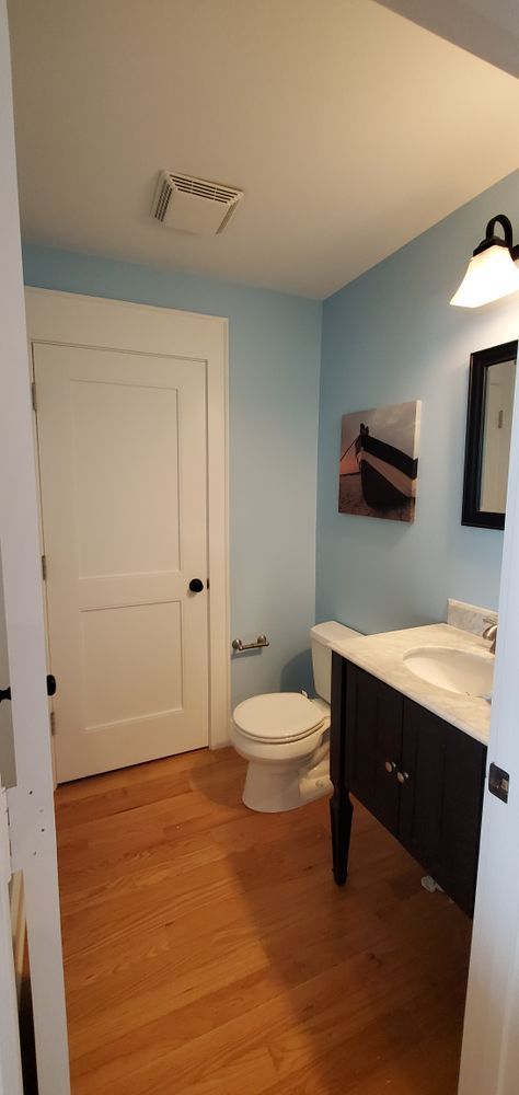 Small bathroom with light blue walls, white door, toilet, vanity, mirror, and wood floor