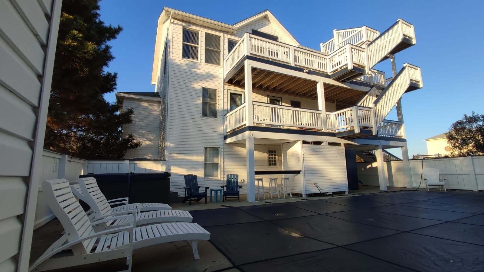 White multi-story house with decks and stairs, viewed from a patio with lounge chairs at sunset