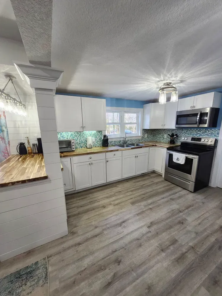 Bright white kitchen with gray wood floors, black appliances, and tile backsplash, viewed from the dining area