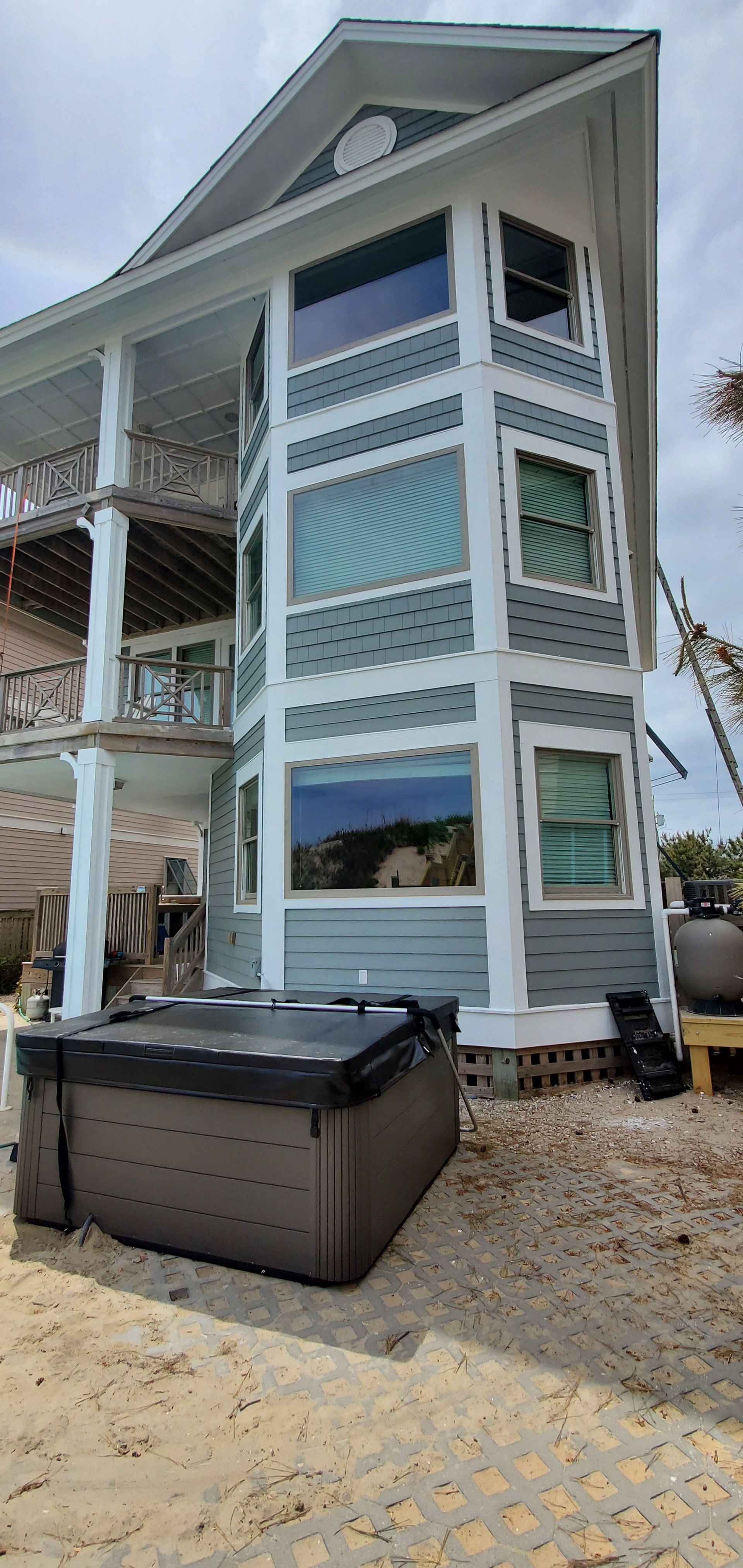 White seaside house with teal shutters and lattice, beside a black hot tub on a pebble patio