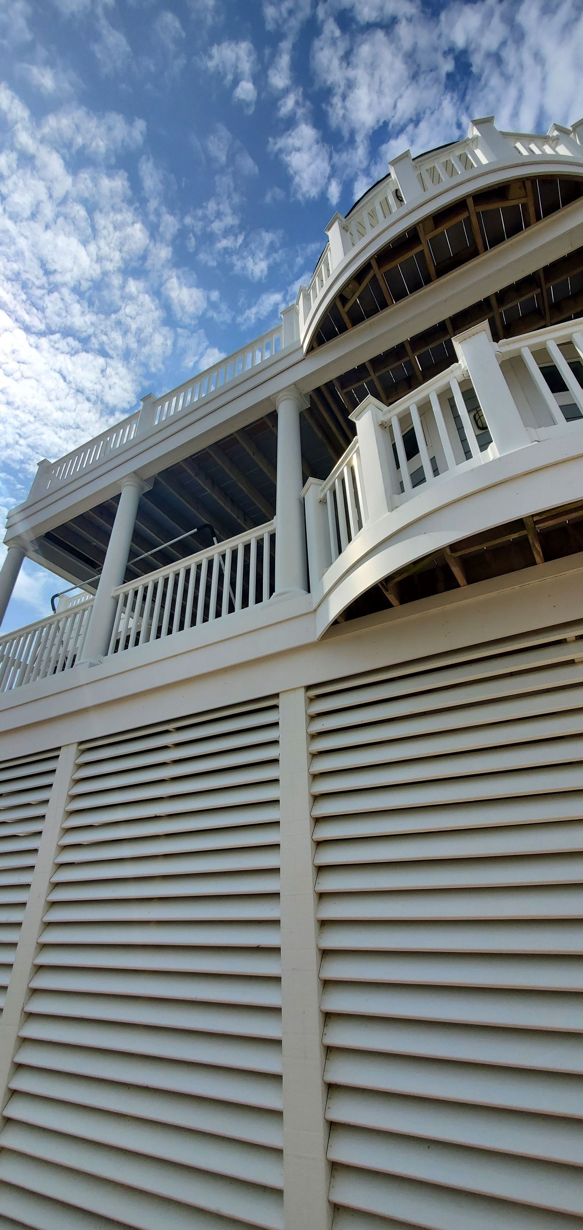 White seaside house with louvered siding and wraparound porch under a cloudy blue sky