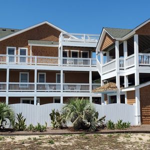 Beachfront condo building with white balconies, brick siding, and a fenced garden under a blue sky