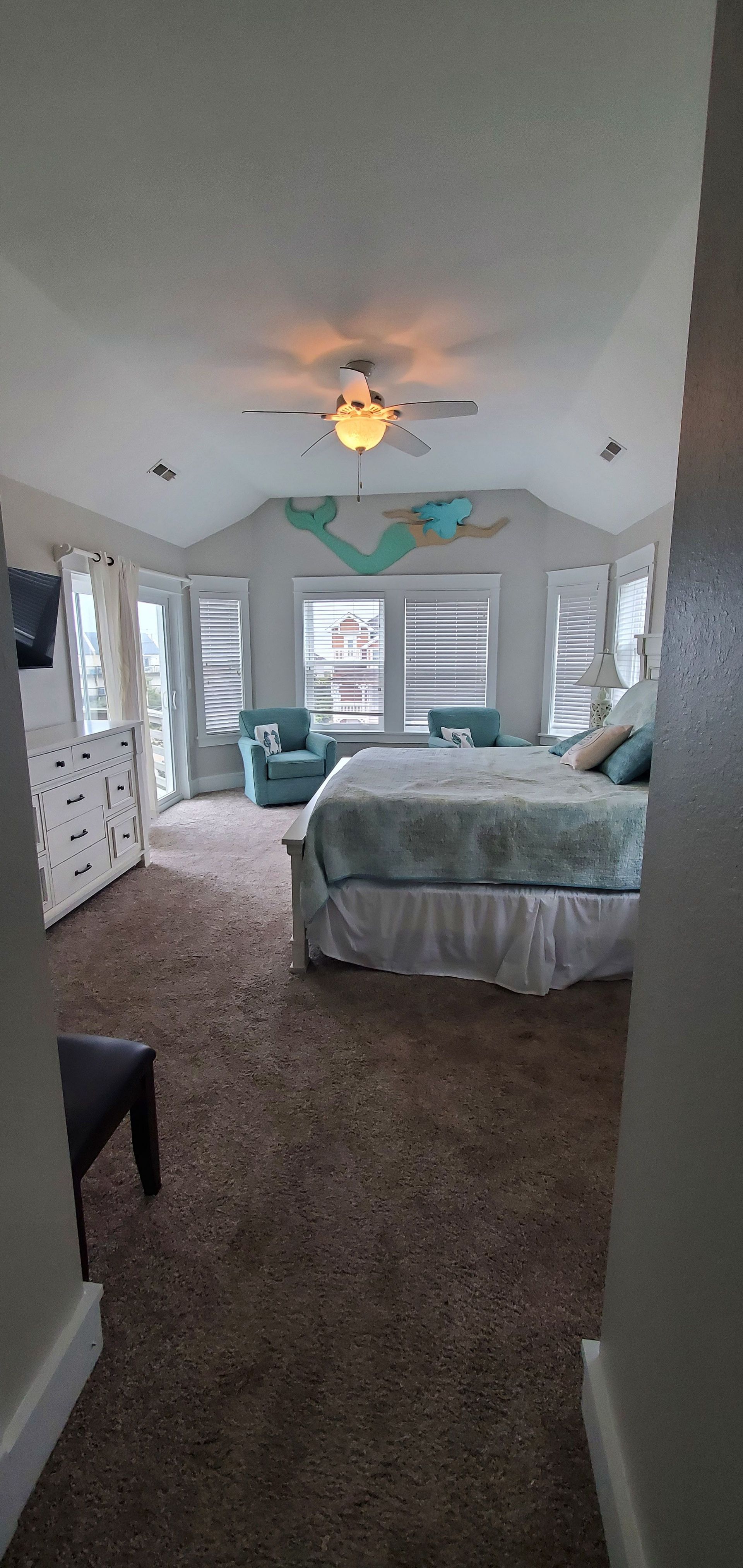 Bright bedroom with two beds, teal chair, ceiling fan, and large windows with white shutters.