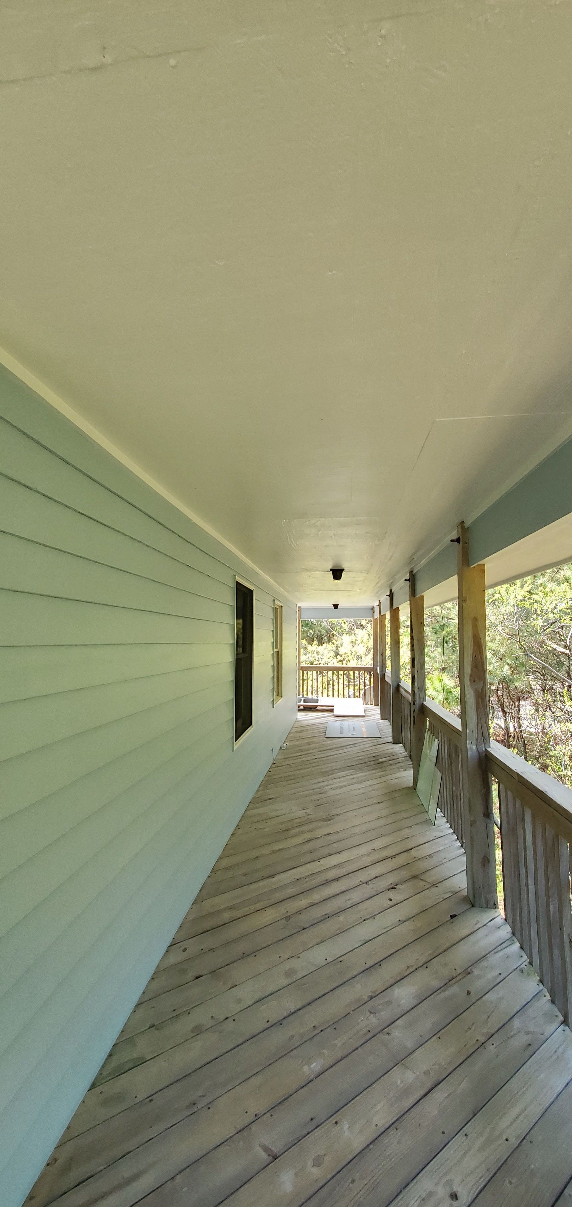 Covered wooden porch with pale green siding, white ceiling, and railing overlooking trees and bright daylight.