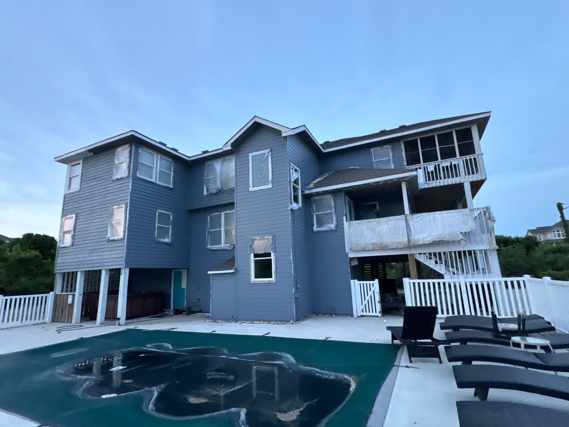 Blue-gray coastal house with a pool and lounge chairs under a cloudy sky