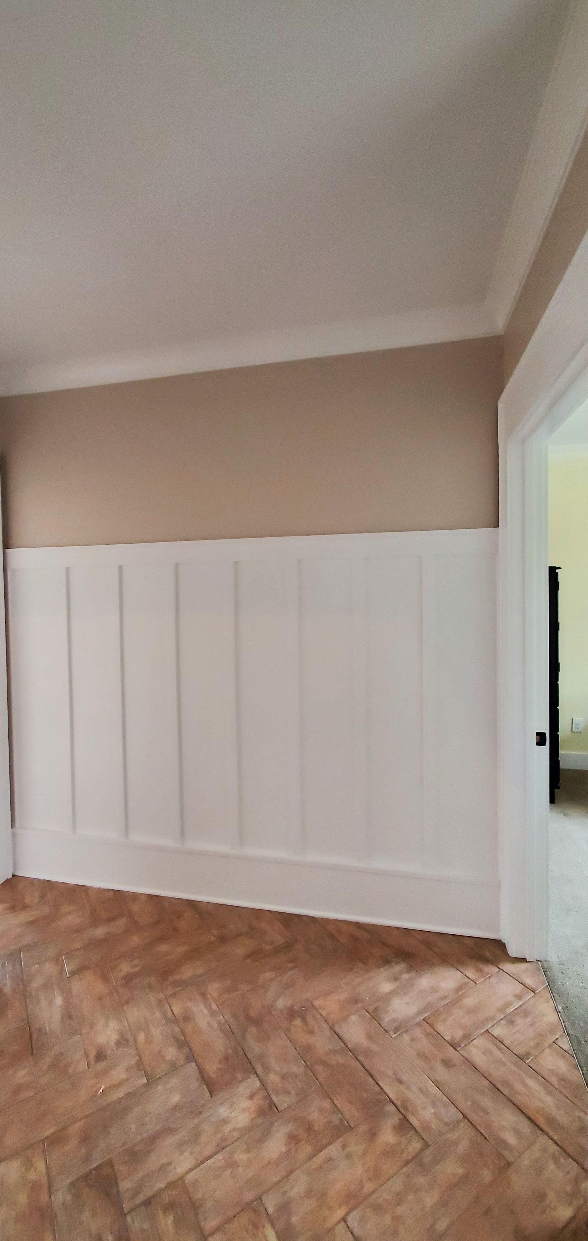 Empty room with white paneled wall, beige upper walls, and herringbone wood floor.