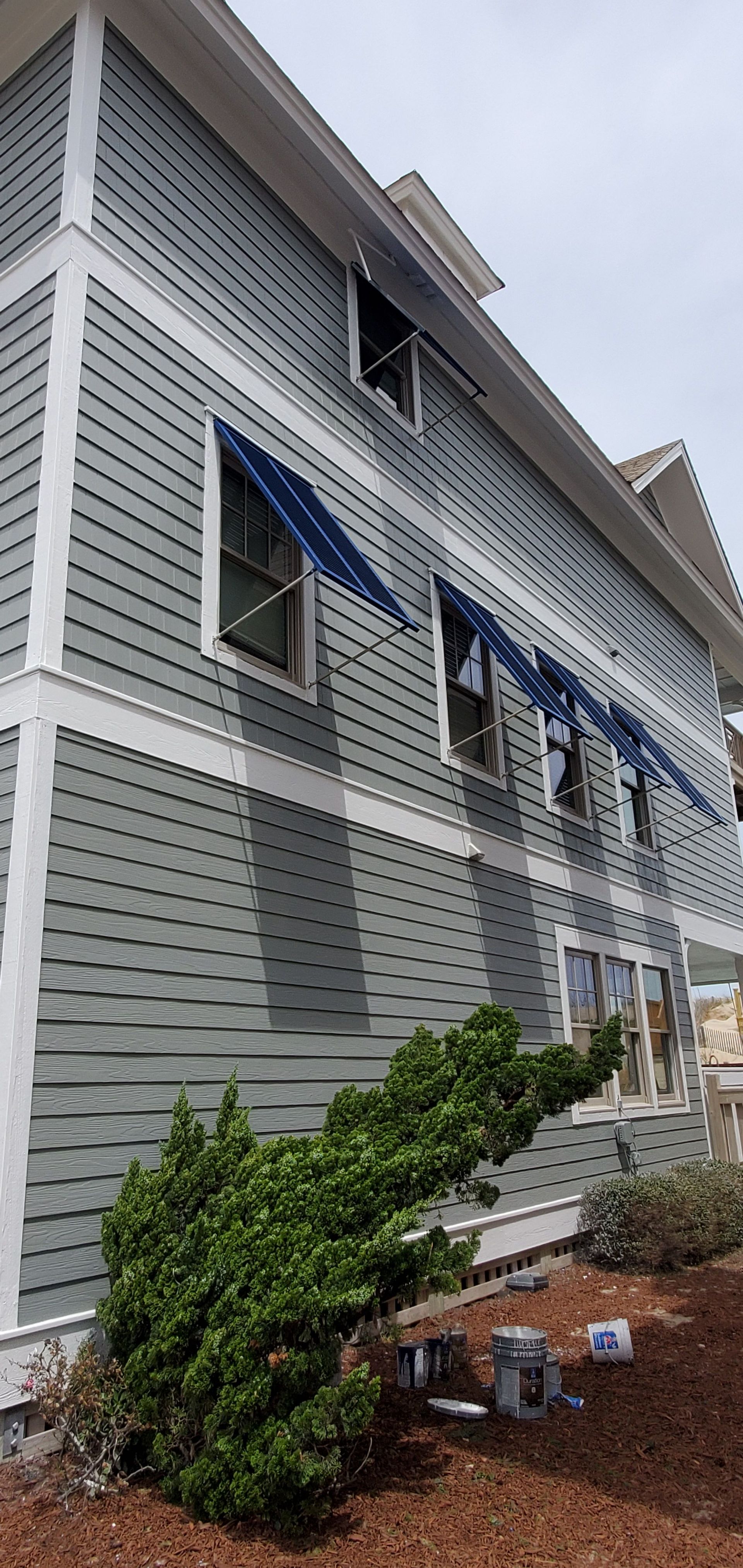 Gray-sided house exterior with windows, white trim, blue awning, and shrubs along a mulch bed