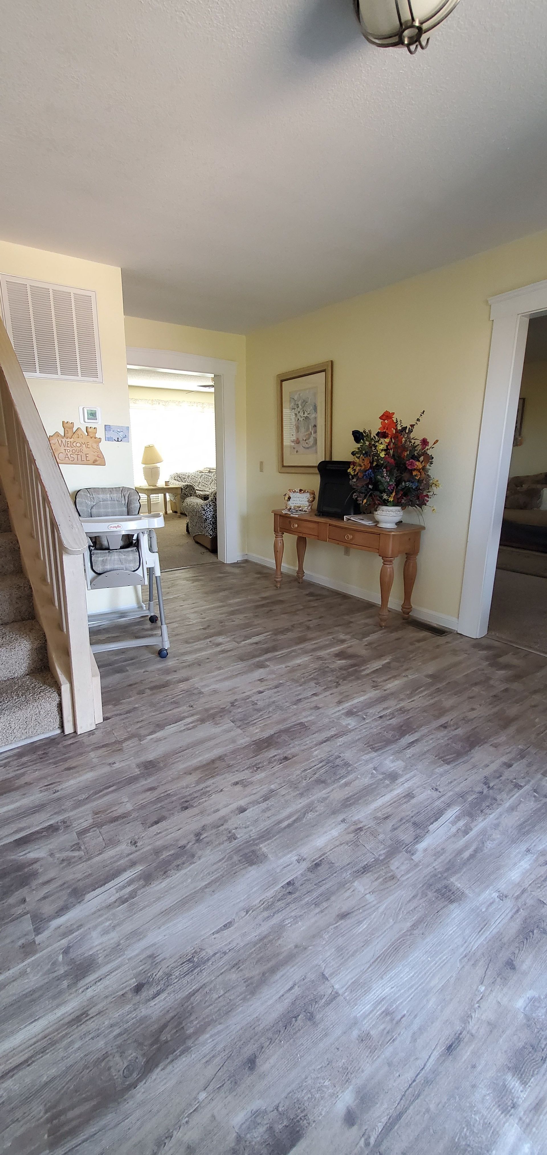 Empty living room with wood-look flooring, a console table with flowers, and an open view to a bright kitchen.