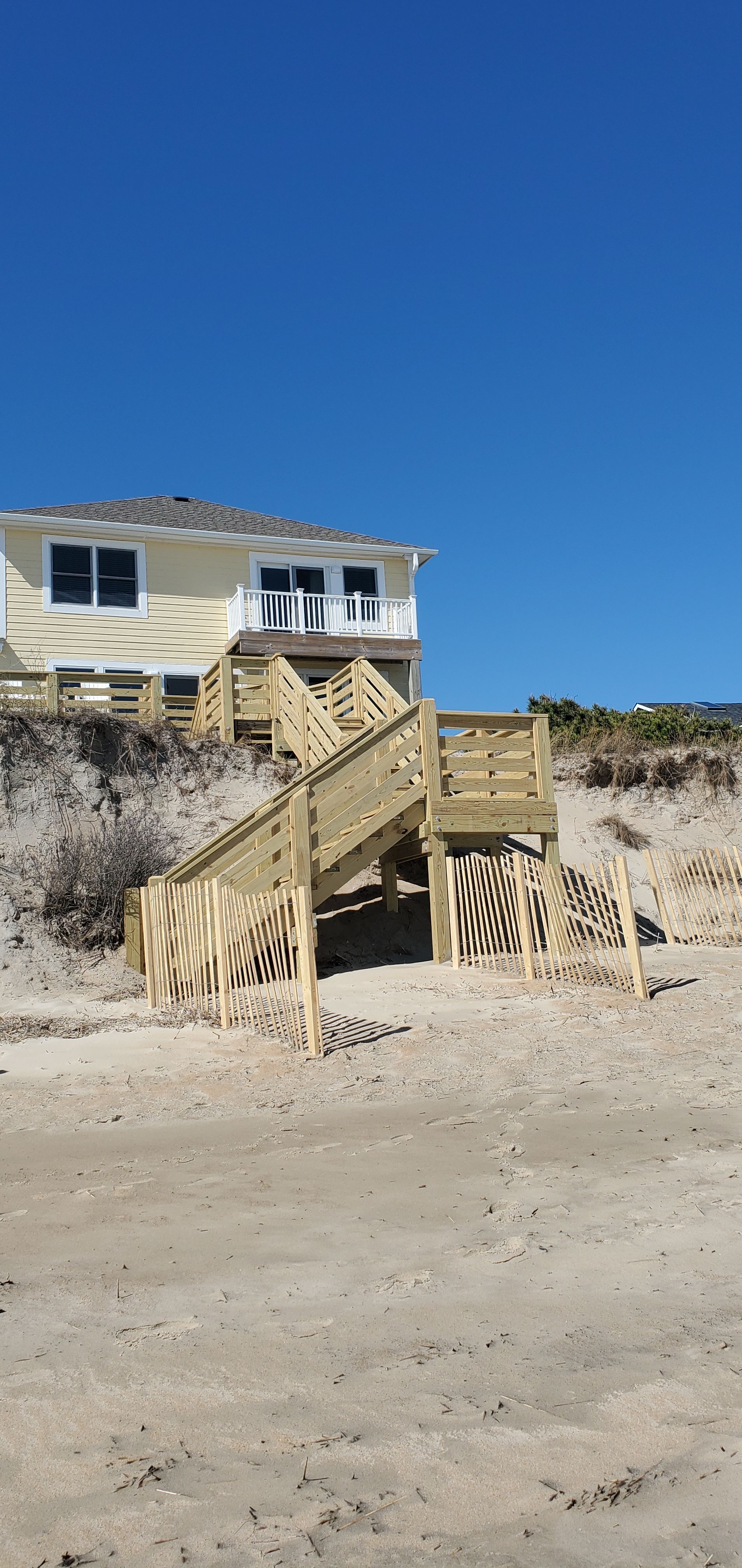 Beach house on stilts above sandy dunes under a clear blue sky.