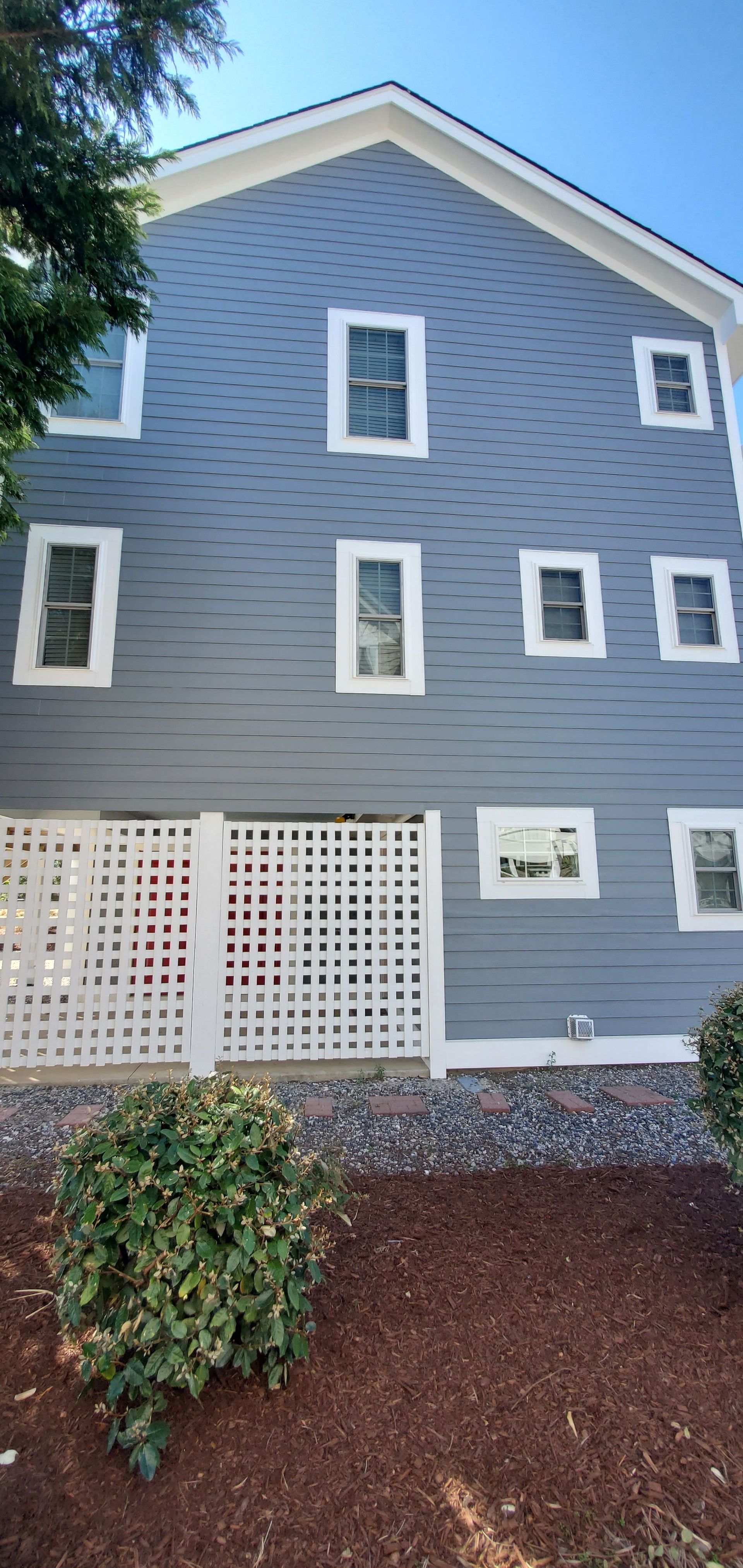 Gray house exterior with white-trimmed windows, white lattice panels, and a small shrub in front.