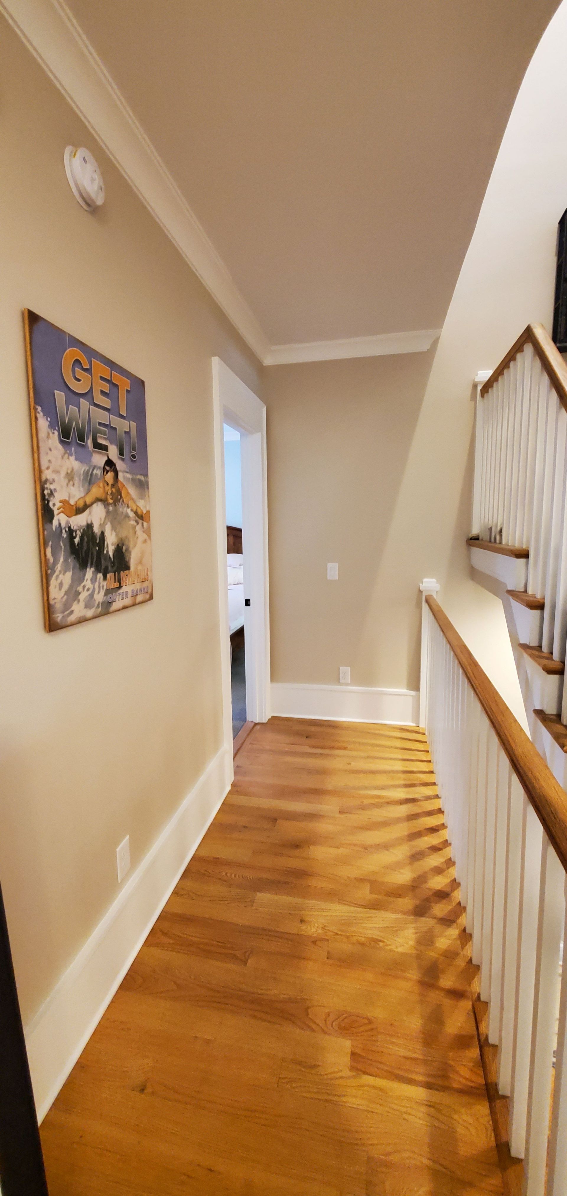 Narrow hallway with tan walls, wooden floor, staircase railing, and framed wall art