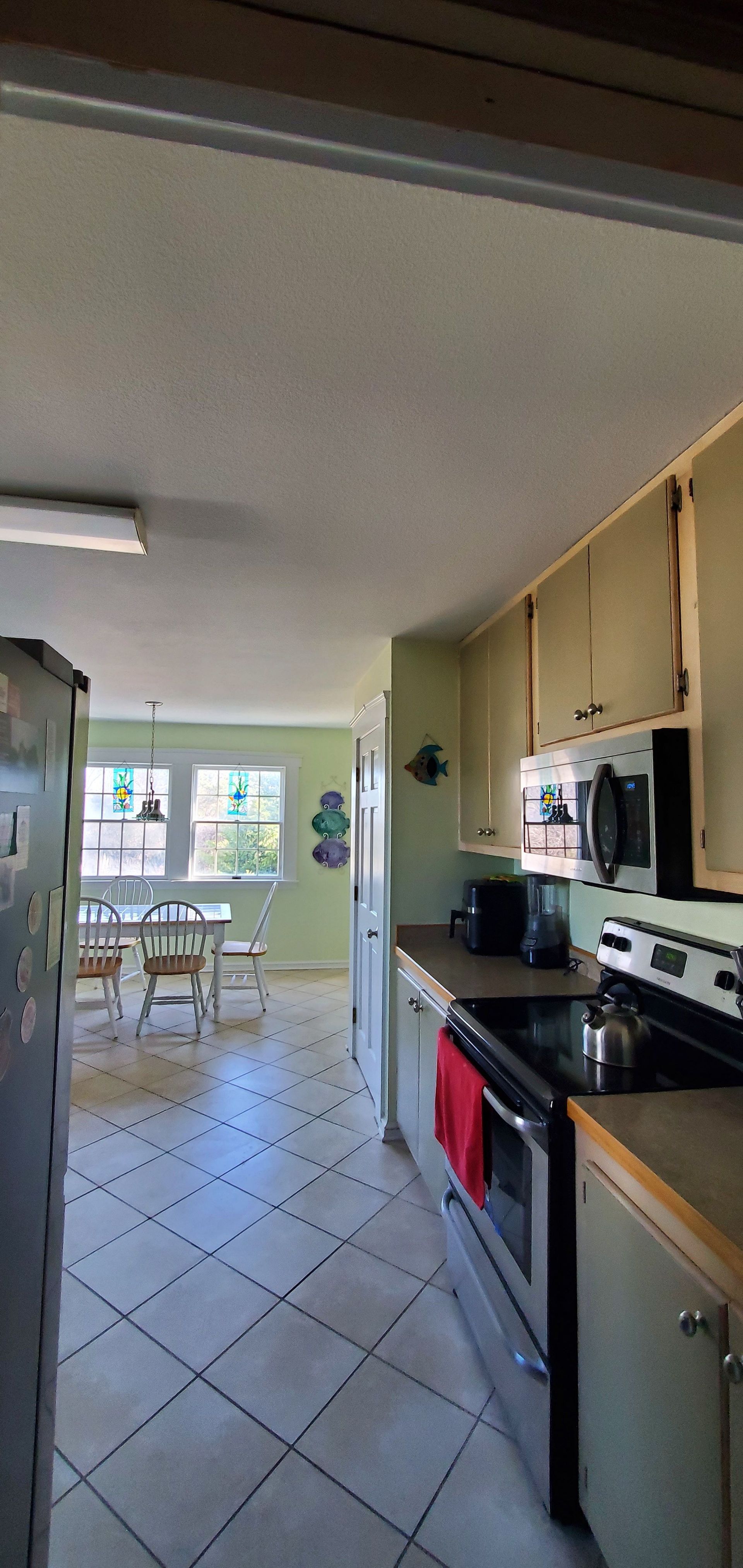 View down a narrow kitchen with tiled floor, wooden cabinets, stove, and dining area by a bright window.