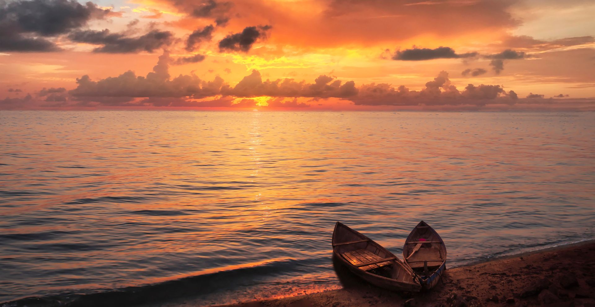 Two small boats on a shore at sunset, with orange clouds reflecting on calm water.