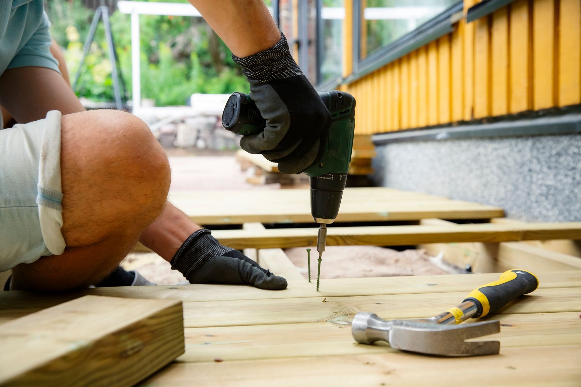 Gloved hands use a power drill to fasten wooden deck boards, with tools nearby.