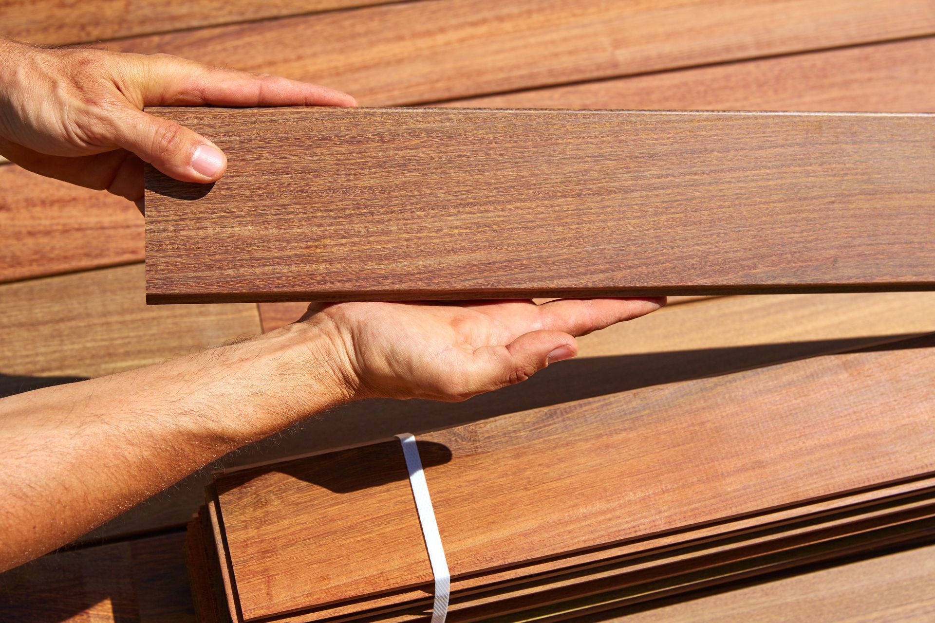 Hands holding a piece of smooth, reddish-brown wood plank against a backdrop of stacked wooden boards.
