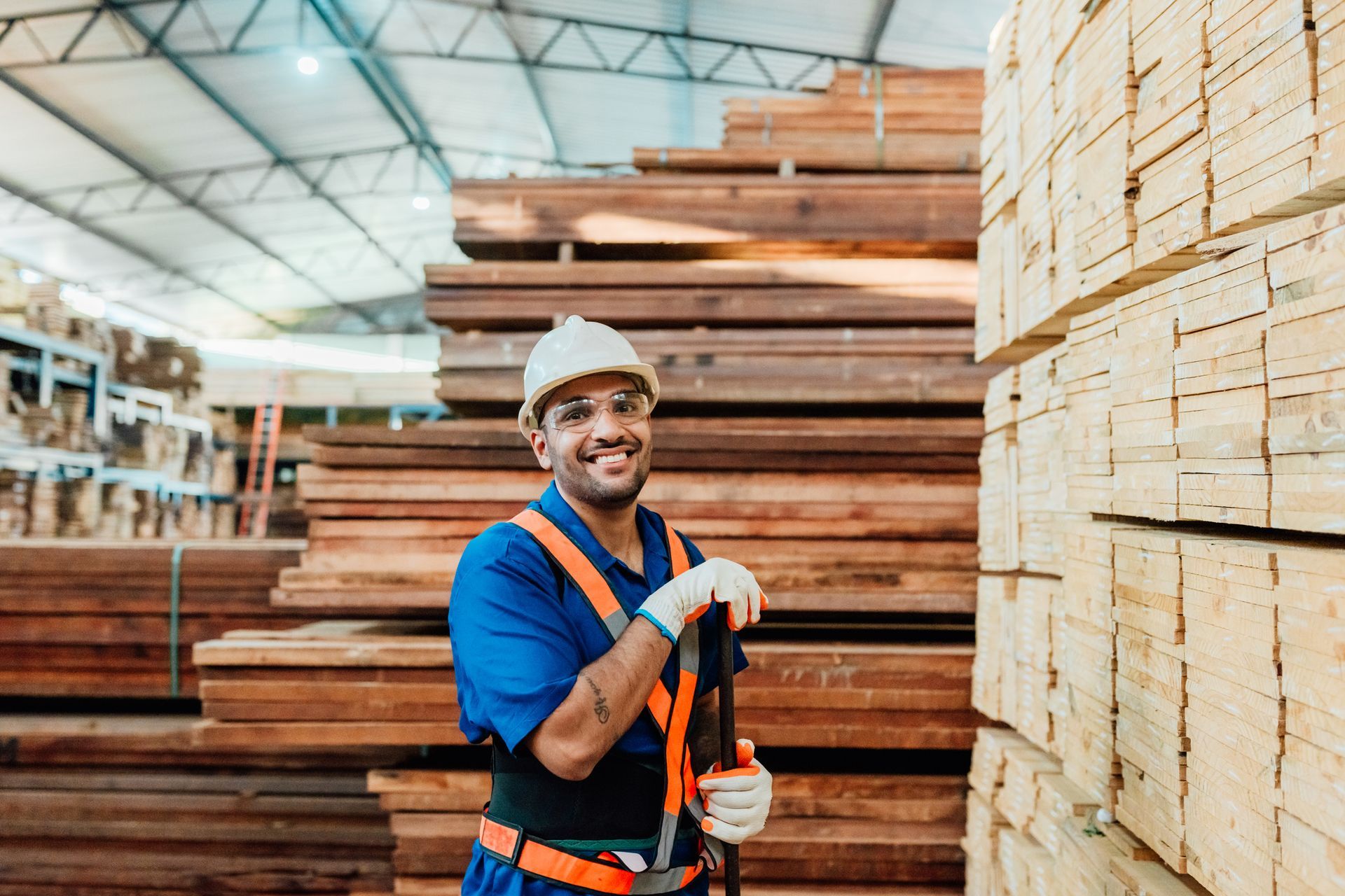 A worker in a lumber warehouse wearing safety gear, holding a tool, with stacks of wood around. A worker in a lumber warehouse wearing safety gear, holding a tool, with stacks of wood around.