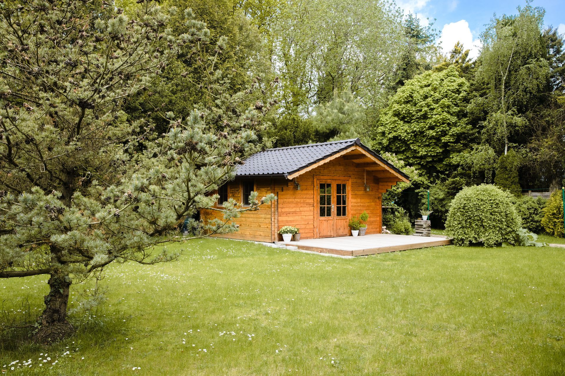 Petite cabane en bois au toit sombre, située dans un jardin verdoyant luxuriant, entourée d'arbres et d'arbustes.