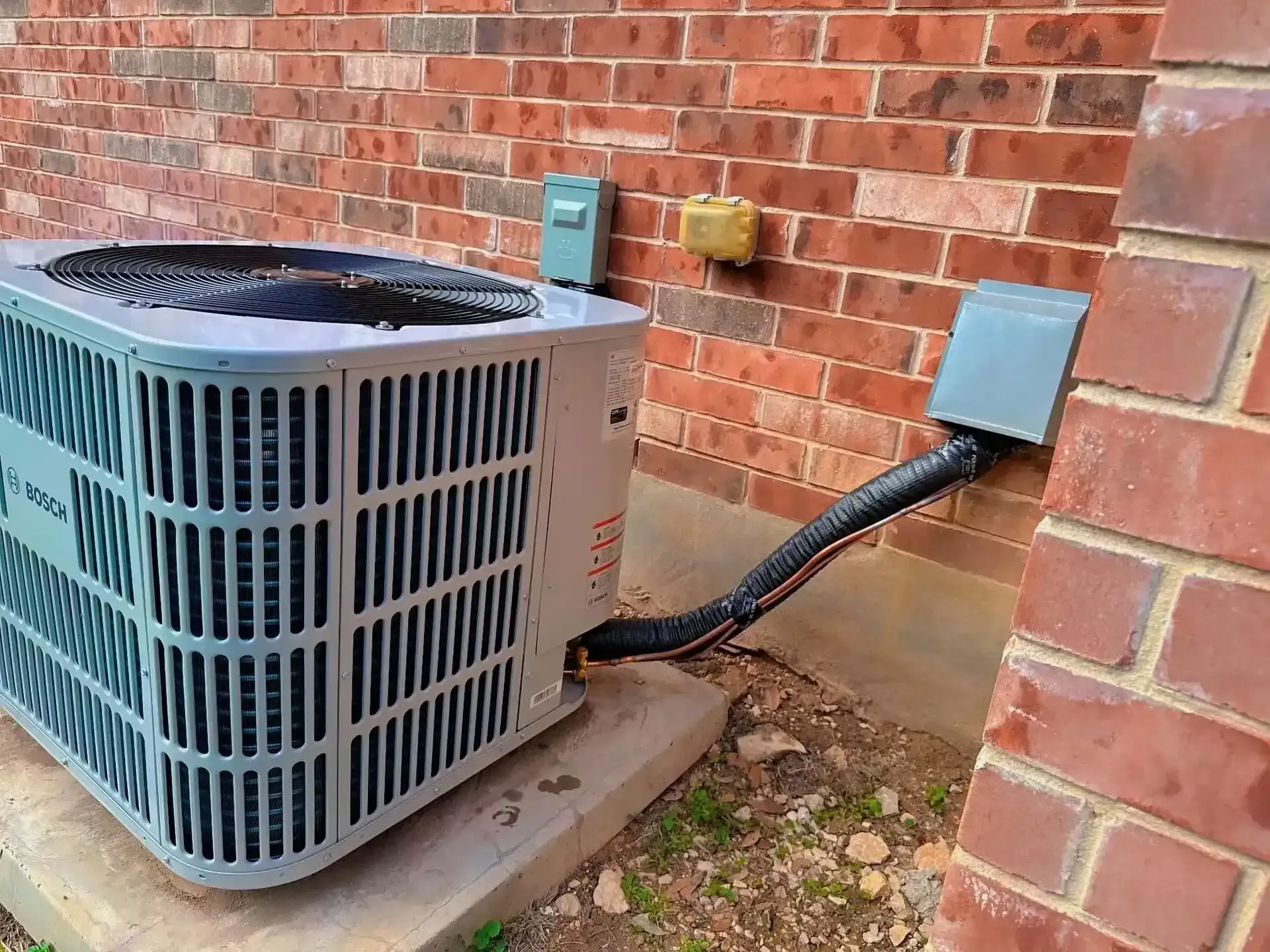 Outdoor air conditioner unit beside a brick house wall, with conduit connected to an electrical box.