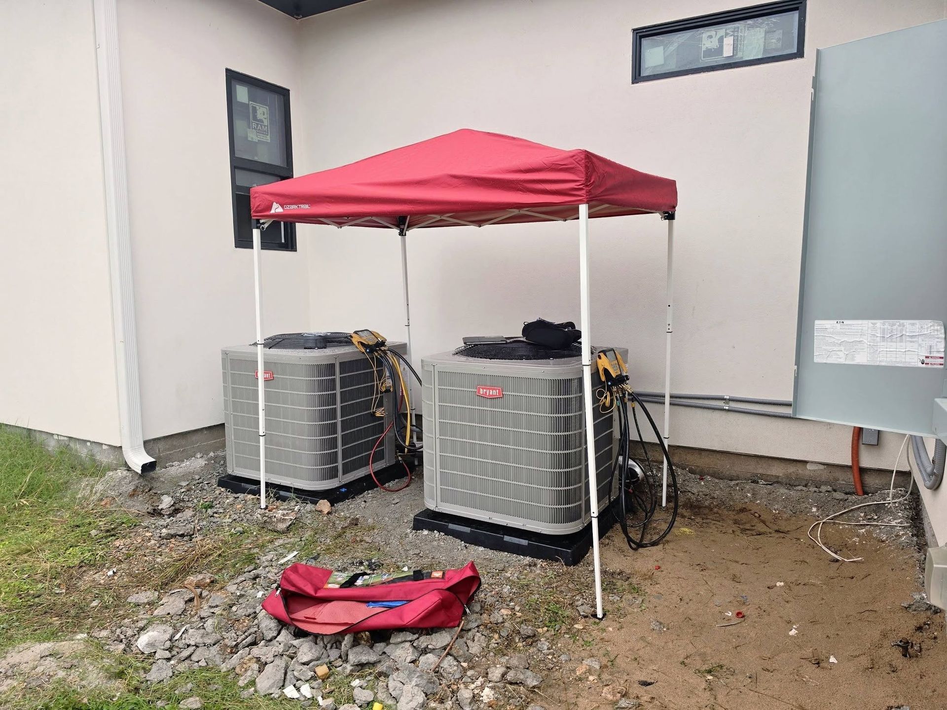 Two outdoor HVAC units under a red canopy beside a building wall, with a red bag on the ground.