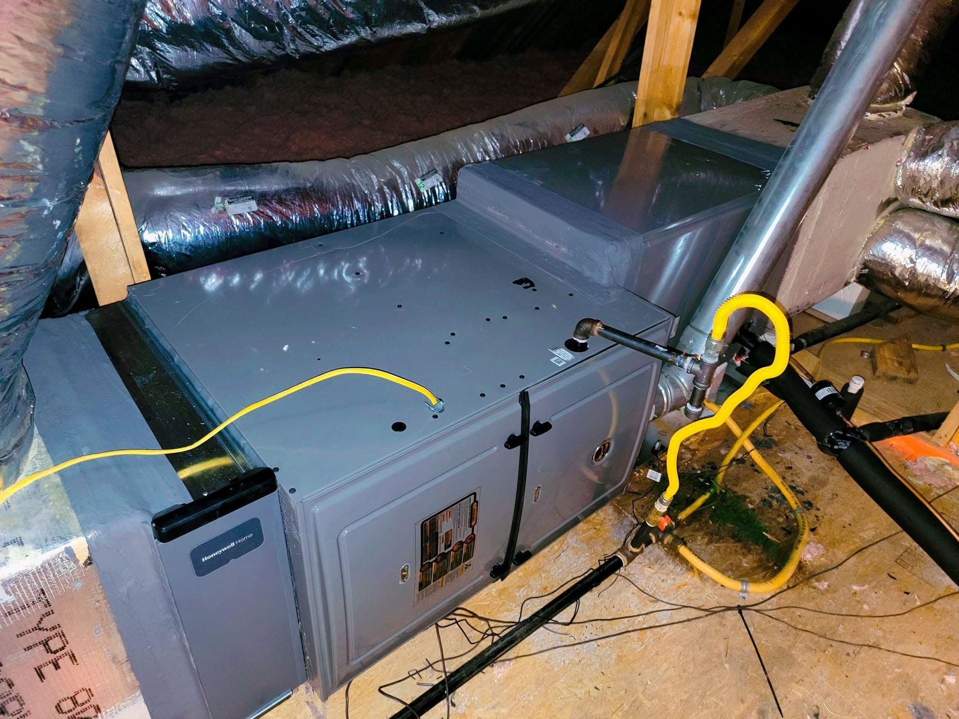 Gray HVAC unit in an attic with yellow cords and exposed ducting.