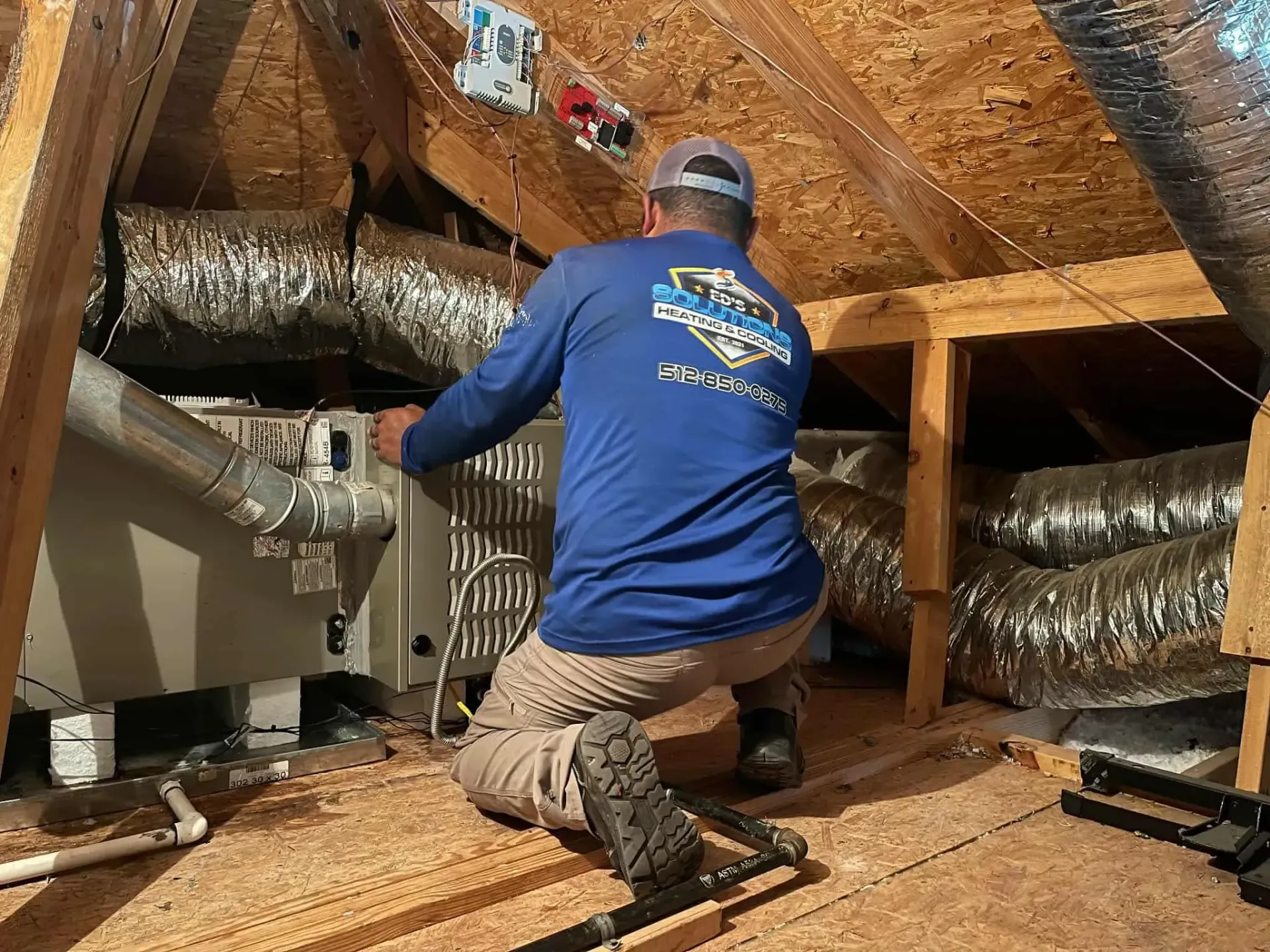 Technician kneeling in an attic, inspecting insulation and ductwork under exposed wooden rafters.