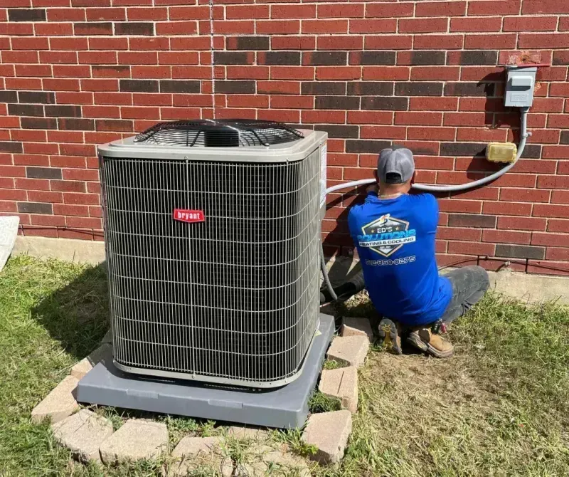 Technician kneeling beside outdoor air conditioner unit against a brick wall