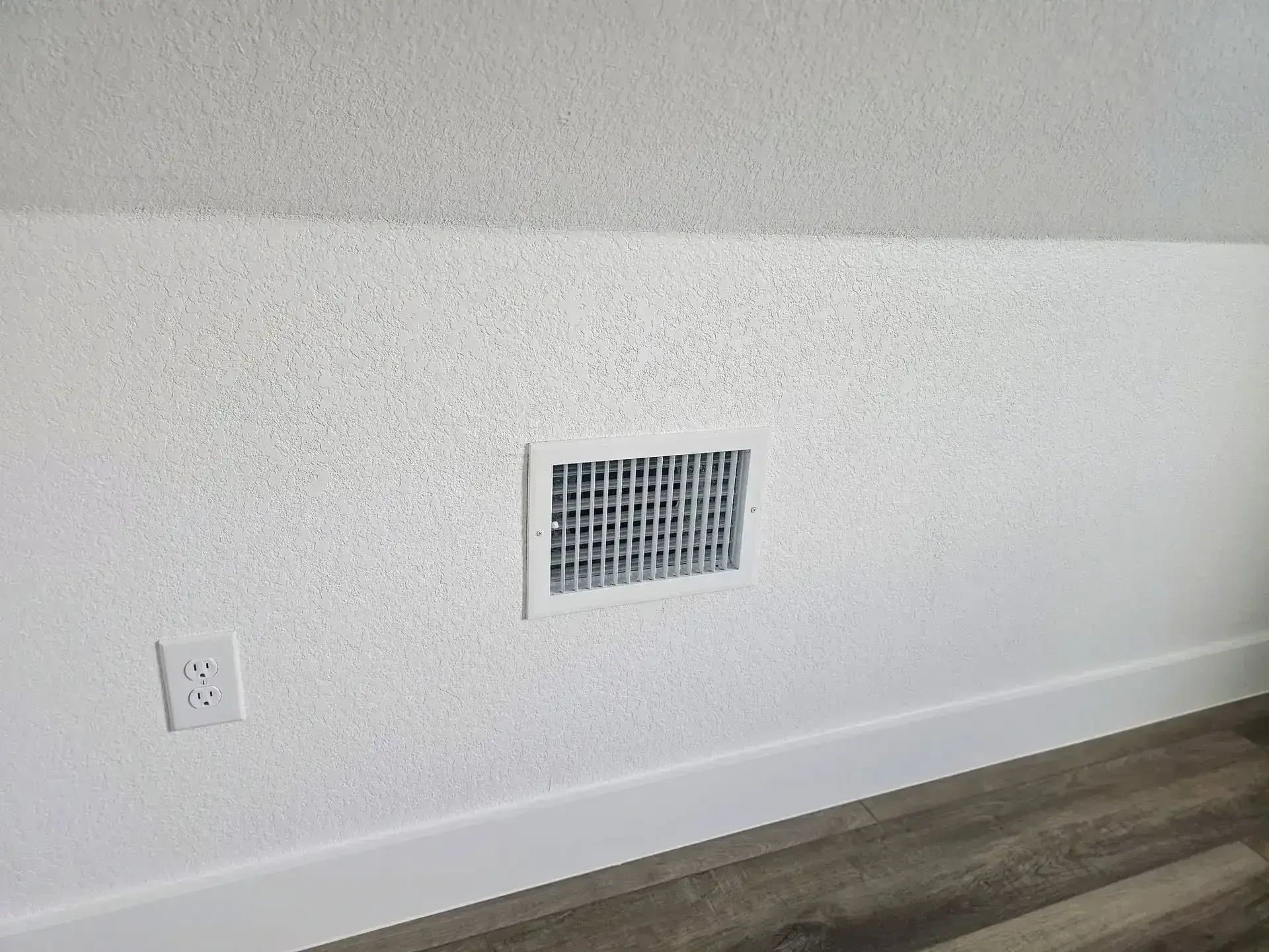 White wall with floor vent and electrical outlet above dark wood flooring.