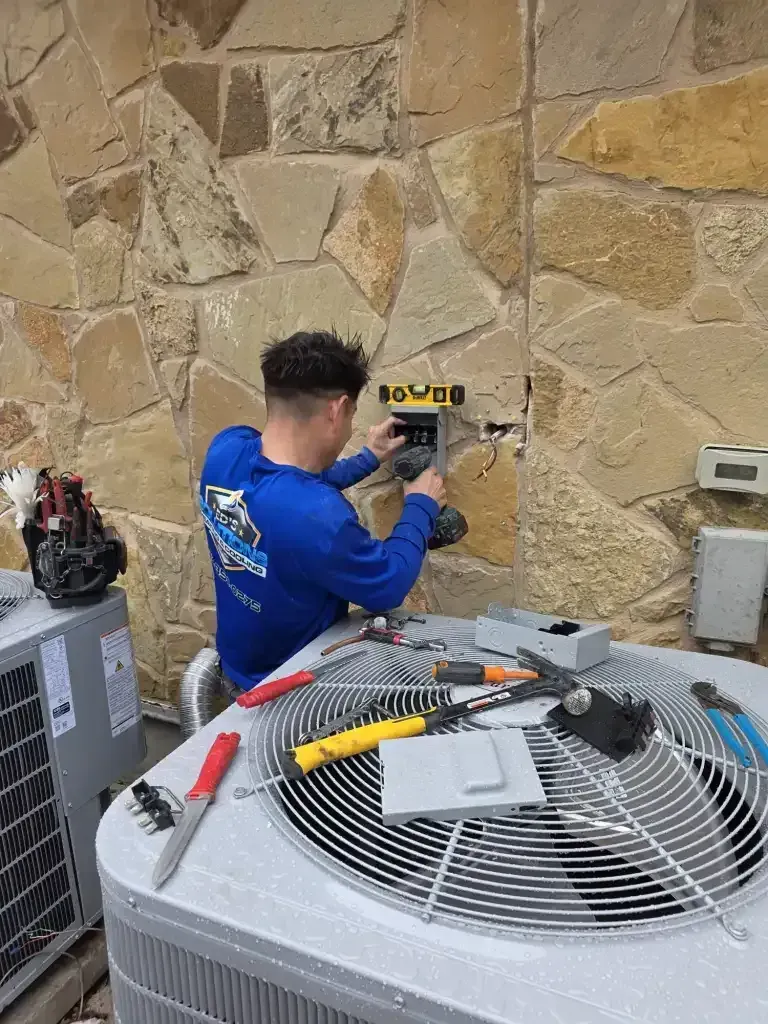 Technician in blue uniform using a drill on a stone wall beside HVAC equipment and tools.