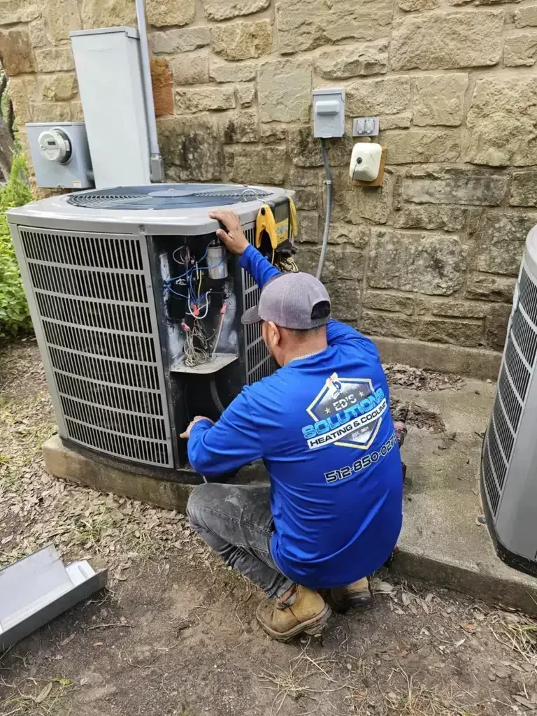 Technician kneels beside an outdoor HVAC unit, repairing it against a stone wall.