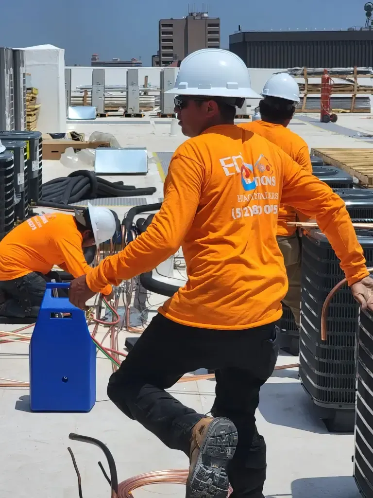 Workers in orange safety shirts and hard hats handling equipment on a rooftop construction site.