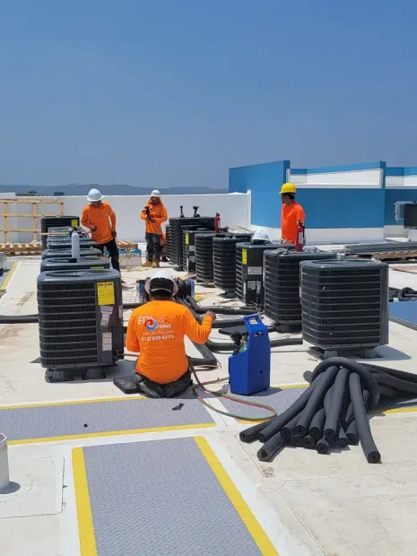 Workers in orange safety gear on a rooftop among black cable coils and equipment under a clear blue sky