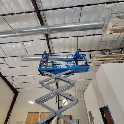 Two workers on a blue scissor lift installing ceiling fixtures in a warehouse-like room.