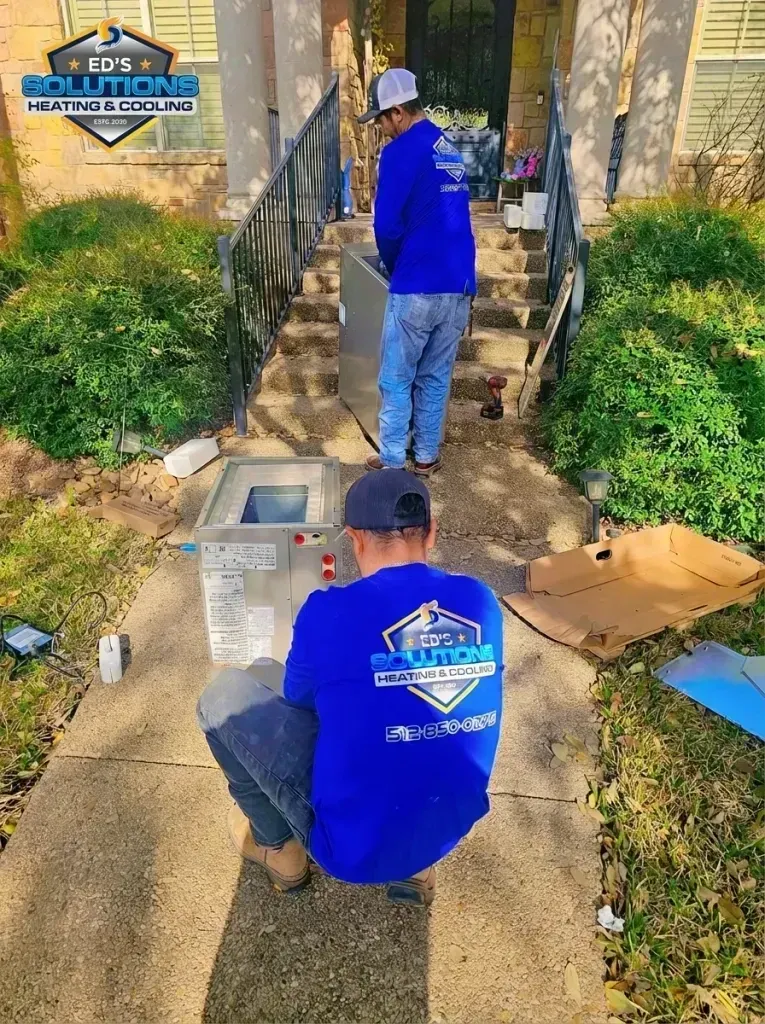 Two workers in blue shirts carry a metal unit up a front walkway to a house