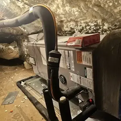 Gas furnace in a dusty crawlspace, with black insulated pipes and warning labels.
