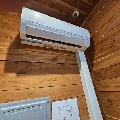 Wall-mounted white air conditioner on a wood-paneled wall above a panel and thermostat.