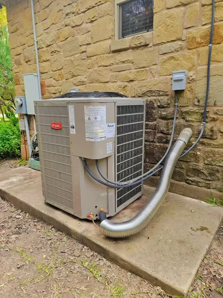 Outdoor HVAC unit beside a stone wall, with metal conduit and piping on a concrete pad.