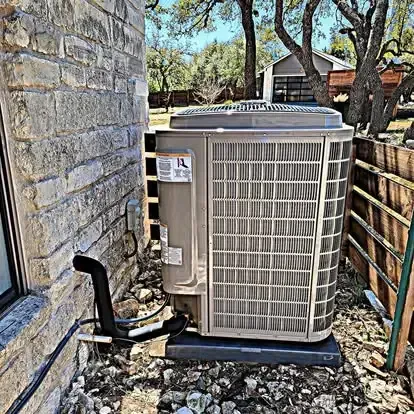 Outdoor HVAC condenser unit beside a stone house wall and wooden fence, with gravel ground and trees nearby