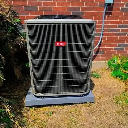 Outdoor air conditioner unit beside a brick wall, with gray grille and red brand badge.