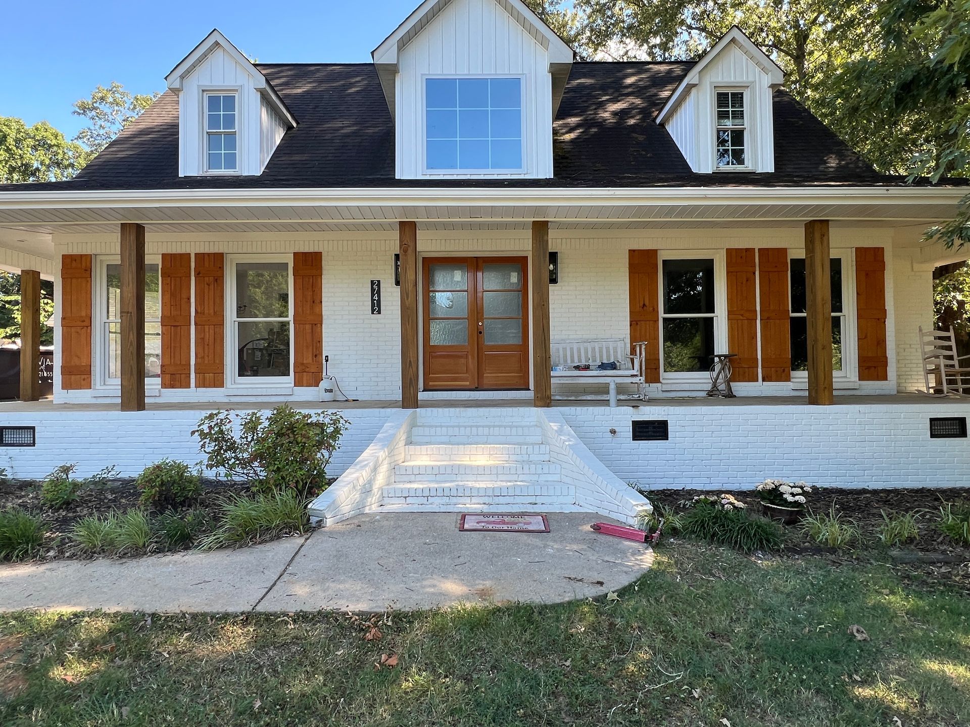 Front view of a white house with a porch, wooden doors, and dormer windows, set behind a walkway.