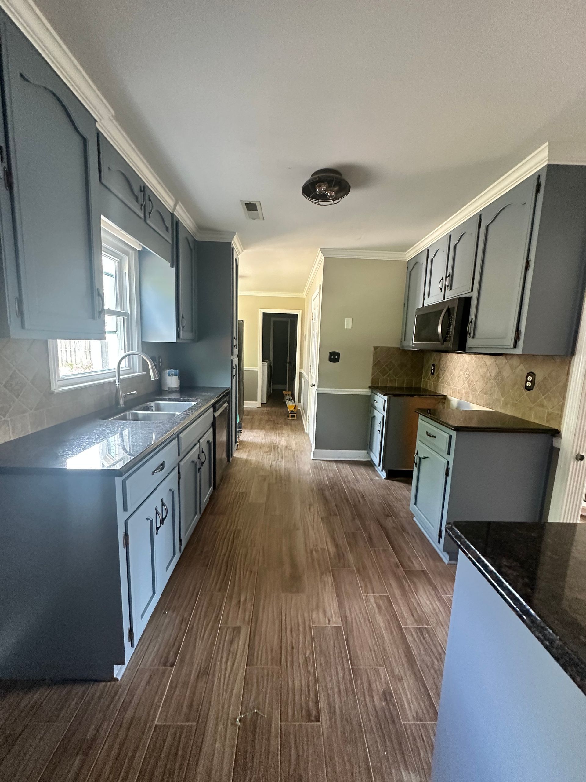 Narrow kitchen with gray cabinets, wood-pattern floor, window light, and a hallway at the far end