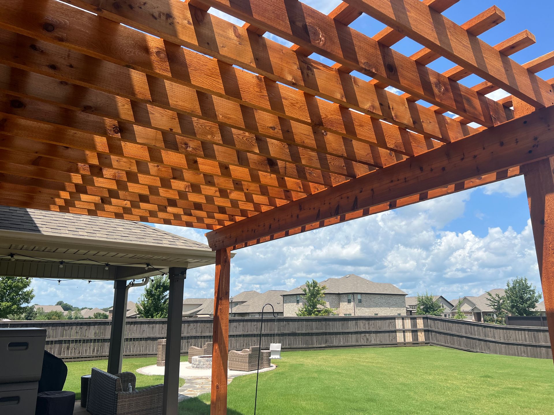 Backyard patio with wooden pergola, grill, lawn, and blue sky over a fenced yard