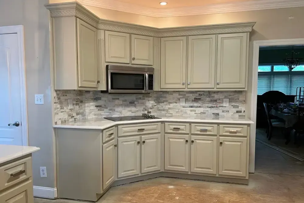Compact kitchen with cream cabinets, mosaic backsplash, microwave, and doorway to a dining area.