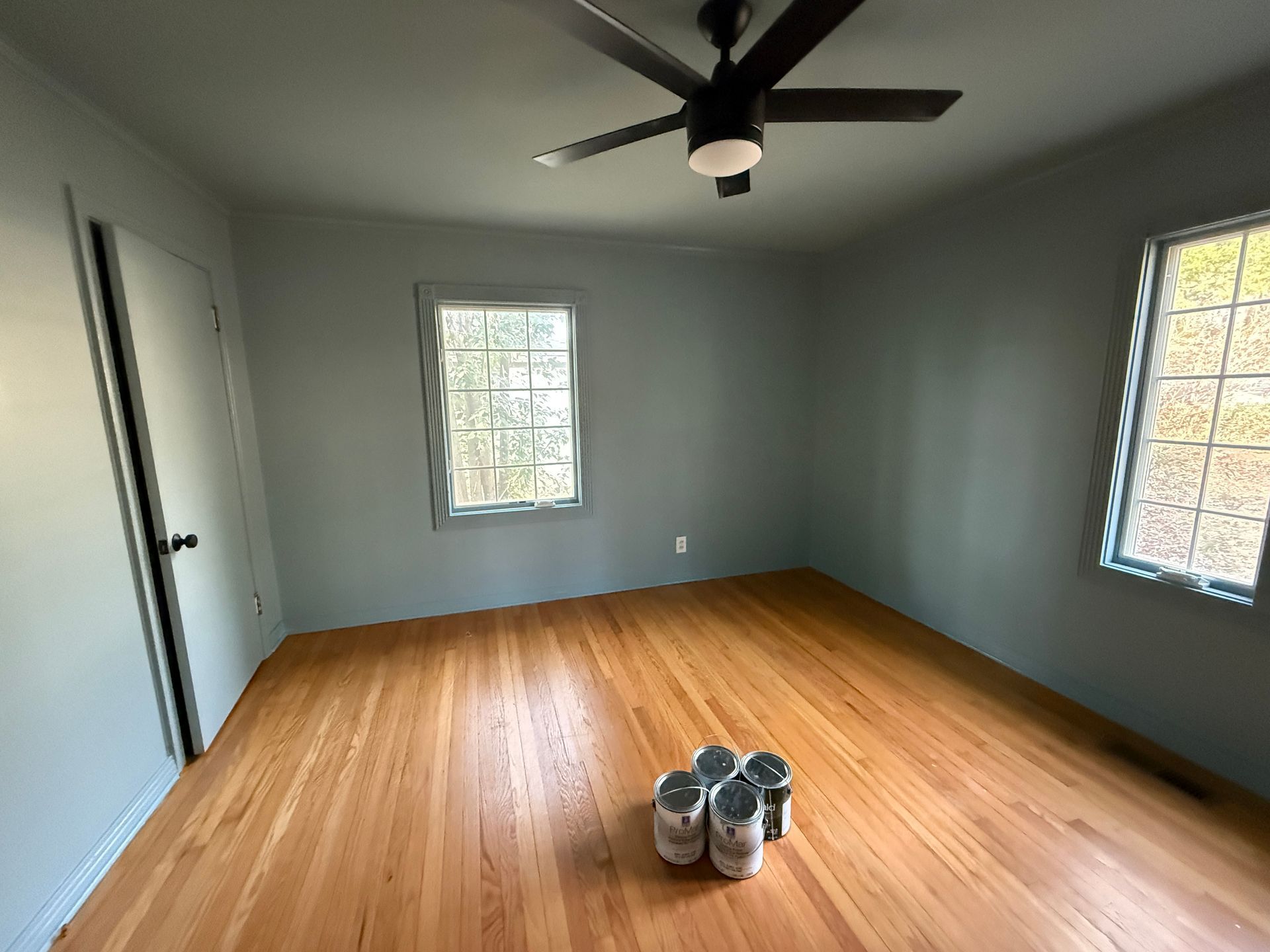 Empty gray bedroom with wood floor, two windows, ceiling fan, and paint cans in the center.