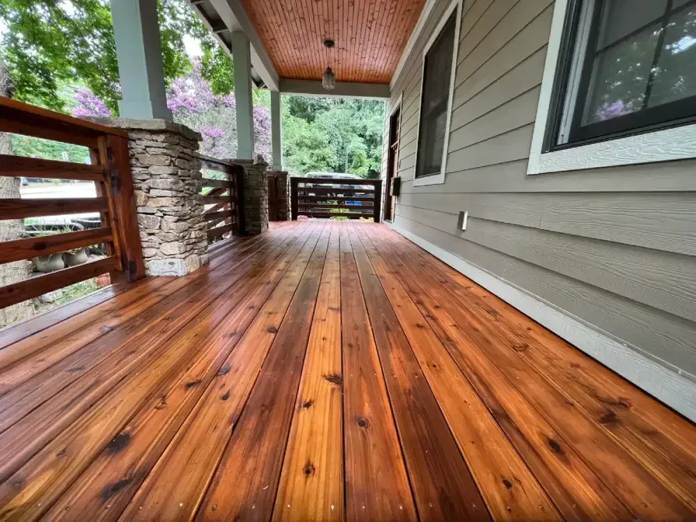 Covered wooden porch with glossy stained floor, stone columns, and railing overlooking greenery.