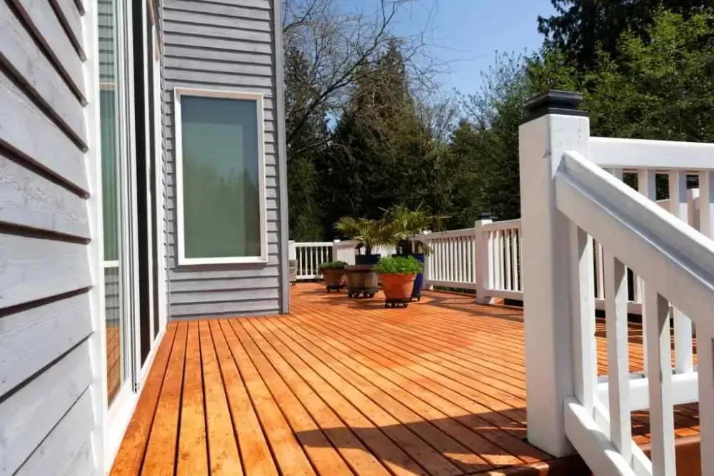 Sunny wooden deck beside a gray house, with white railings and potted plants.